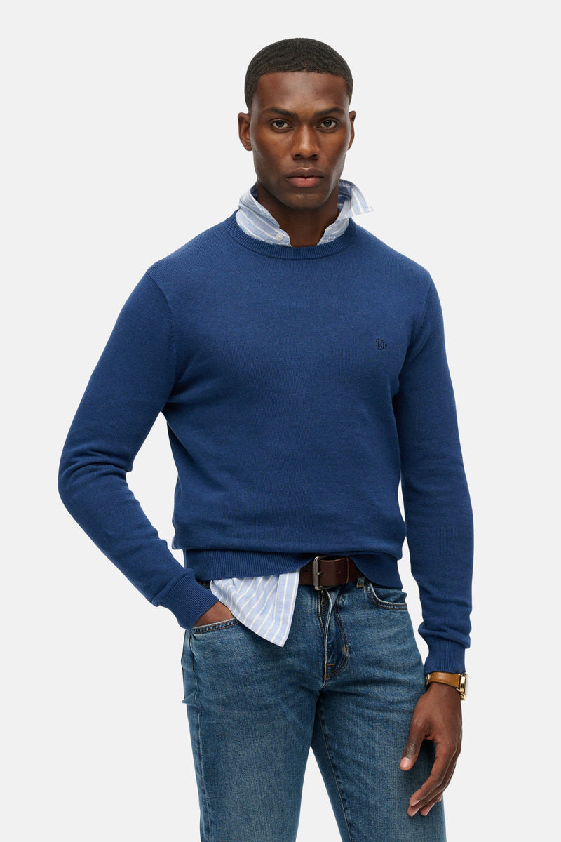 Pull col rond bleu de SUPERDRY porté sur une chemise rayée, avec un jean et une montre en cuir.