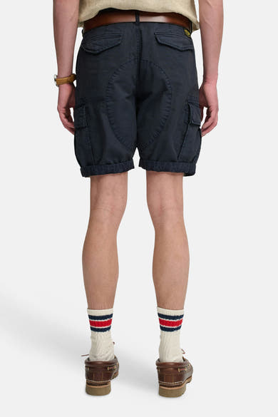 Korte donkerblauwe cargobroek van SUPERDRY, van achteren gezien, met zakken, een gele patch en een omgeslagen zoom, gedragen met een bruine riem en gestreepte sokken.
