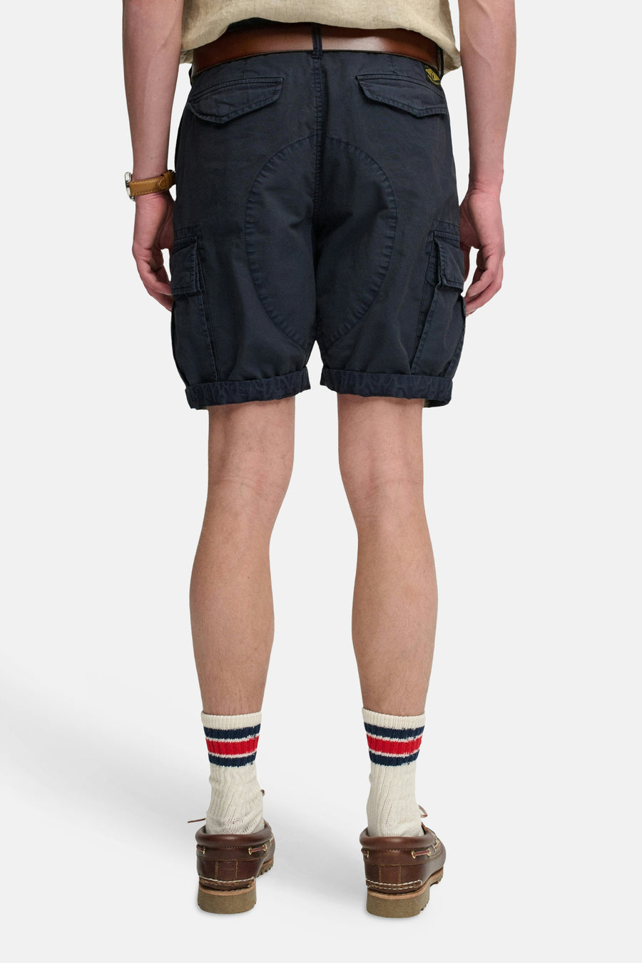Korte donkerblauwe cargobroek van SUPERDRY, van achteren gezien, met zakken, een gele patch en een omgeslagen zoom, gedragen met een bruine riem en gestreepte sokken.