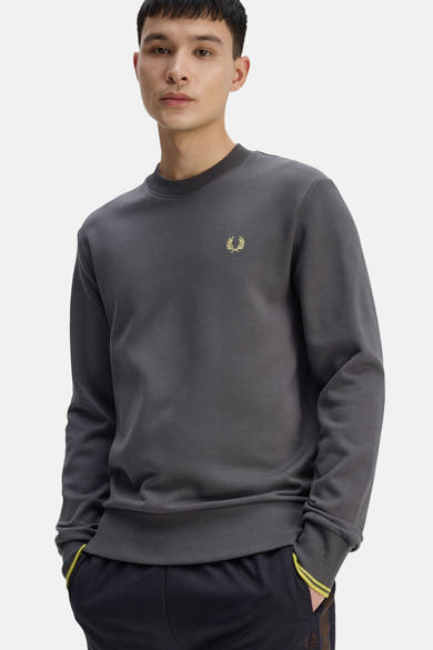 Sweat col rond gris de Fred Perry, orné d'un logo doré sur la poitrine, porté avec un pantalon noir.