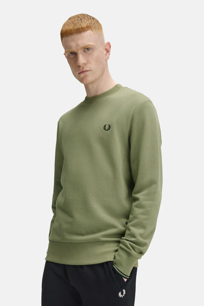 Olivengroene trui van Fred Perry, met het merklogoogd op de borst en gedragen met een zwarte broek.