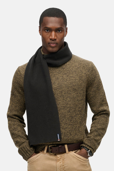 Écharpe noire par Superdry, portée avec un pull texturé couleur olive et un pantalon beige.
