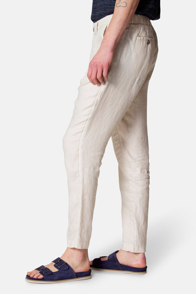 Pantalon de costume beige de Le Fabuleux Marcel de Bruxelles, porté avec un t-shirt bleu marine et des sandales bleues.