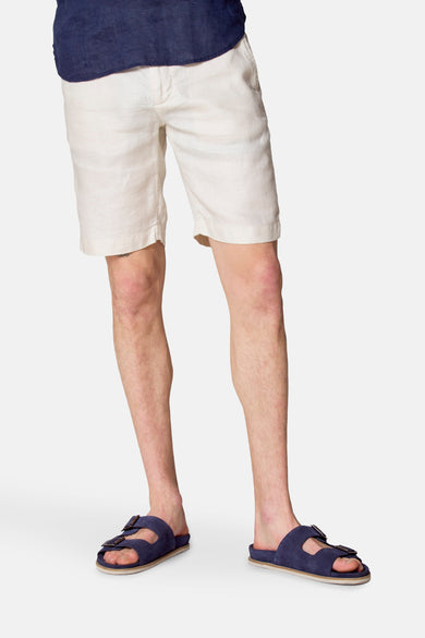 Short beige en lin de Le Fabuleux Marcel de Bruxelles, porté avec une chemise bleu marine et des sandales bleues.