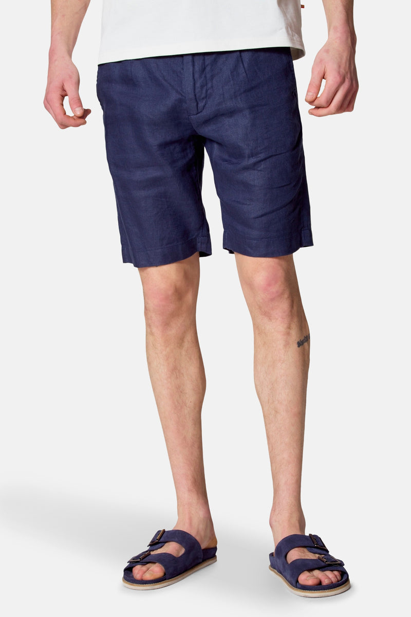 Short bleu en lin, de Le Fabuleux Marcel de Bruxelles, porté avec un t-shirt blanc et des sandales bleues.