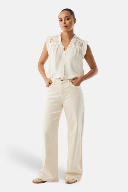 Blouse blanche sans manches de MEXX, avec détails ajourés et volants aux épaules, portée avec un pantalon crème.
