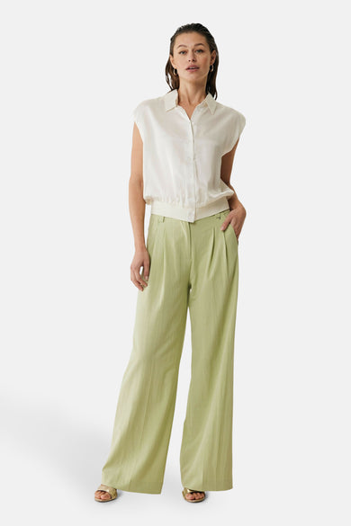 Blouse écru sans manches de MEXX, portée avec un pantalon vert et des sandales dorées.