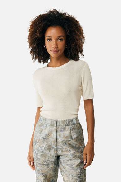 Top écru en maille de MEXX, porté avec un pantalon à motif abstrait, cheveux afro bouclés et bijoux dorés.
