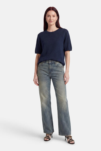 Pull bleu à col rond de MEXX, à manches courtes, porté avec un jean et des sandales à talons.
