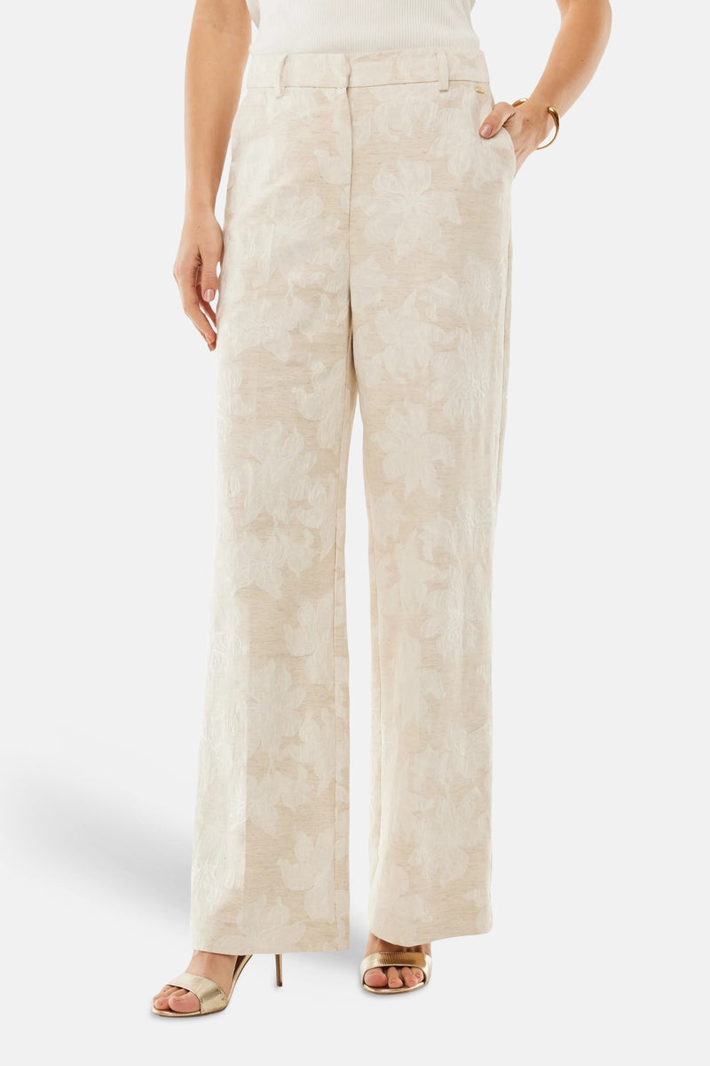 Pantalon beige de MEXX, avec un motif floral texturé, coupe droite et jambes larges, porté avec des chaussures à talons dorées.
