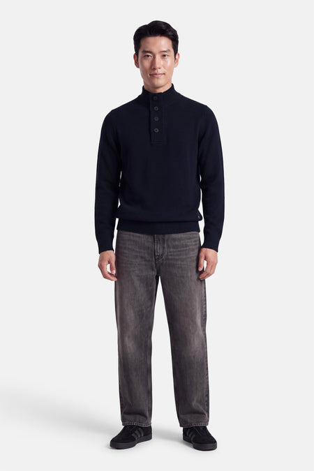Pull col roulant bleu marine de BARBOUR CLASSIC, à boutons discrets, tricoté, porté avec jeans gris foncé et baskets noires, vue frontale.