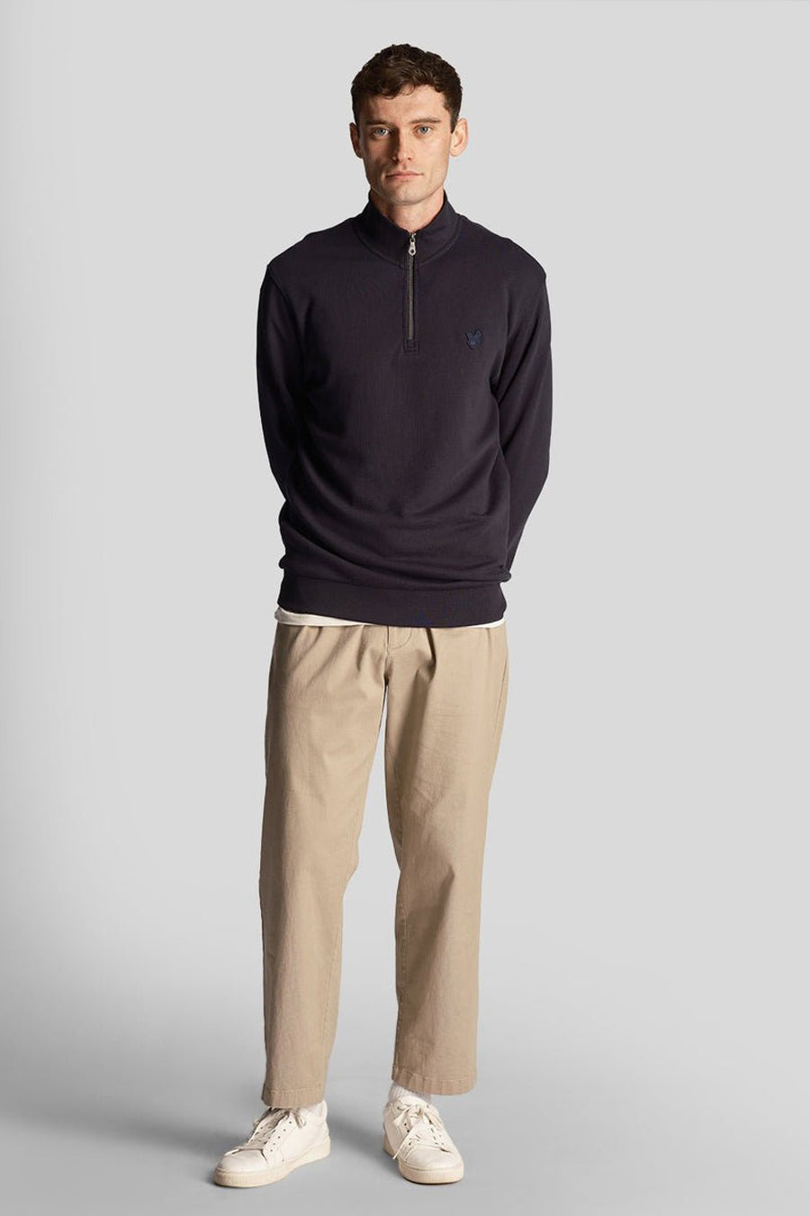 Lyle & Scott blauwe sweater met opstaande kraag, gedragen met een beige broek en witte sneakers.
