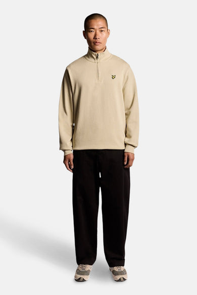 Sweat beige à col zippé de LYLE & SCOTT, porté avec un pantalon noir ample et des baskets.