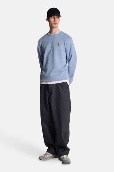 Sweat bleu de LYLE & SCOTT, porté avec un t-shirt blanc, un pantalon ample noir et des baskets grises.