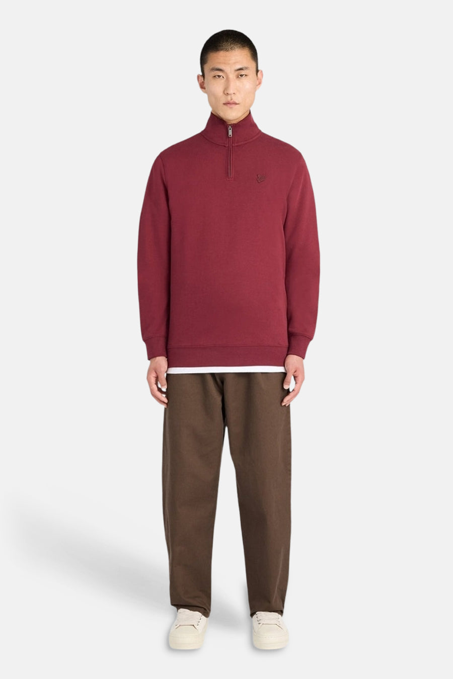Burgundy half-zip sweatshirt with ribbed cuffs and hem, featuring small embroidered logo on chest, worn by model in front view with brown pants and white sneakers.