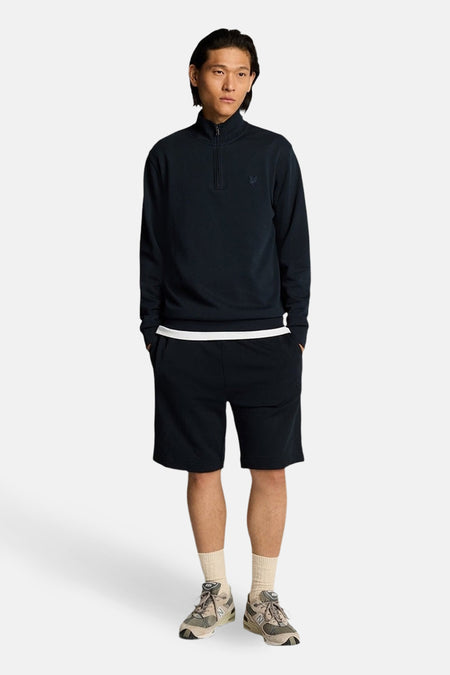 Blauwe sweater met opstaande kraag van LYLE & SCOTT, gedragen met een zwarte korte broek en sneakers aan de voeten.
