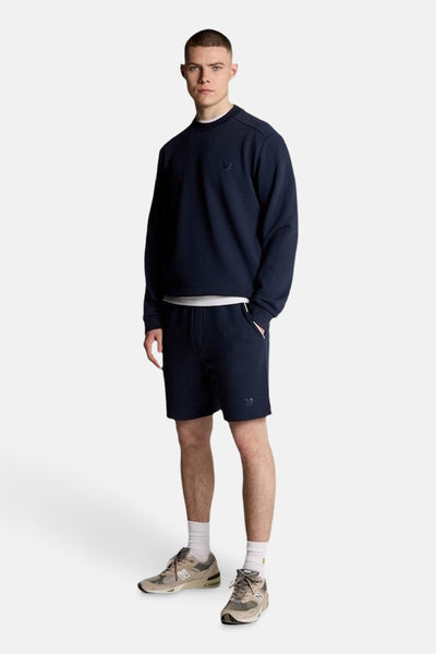 Sweat bleu de LYLE & SCOTT, porté avec un t-shirt blanc et un short de survêtement bleu marine.