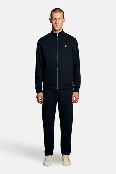 Gilet bleu par LYLE & SCOTT, avec fermeture éclair sur fond uni, porté avec un pantalon de survêtement et des baskets claires.