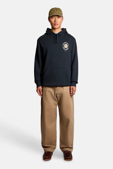 Sweat à capuche bleu de LYLE & SCOTT avec motif floral et texte sur la poitrine, porté avec un pantalon beige et une casquette kaki.