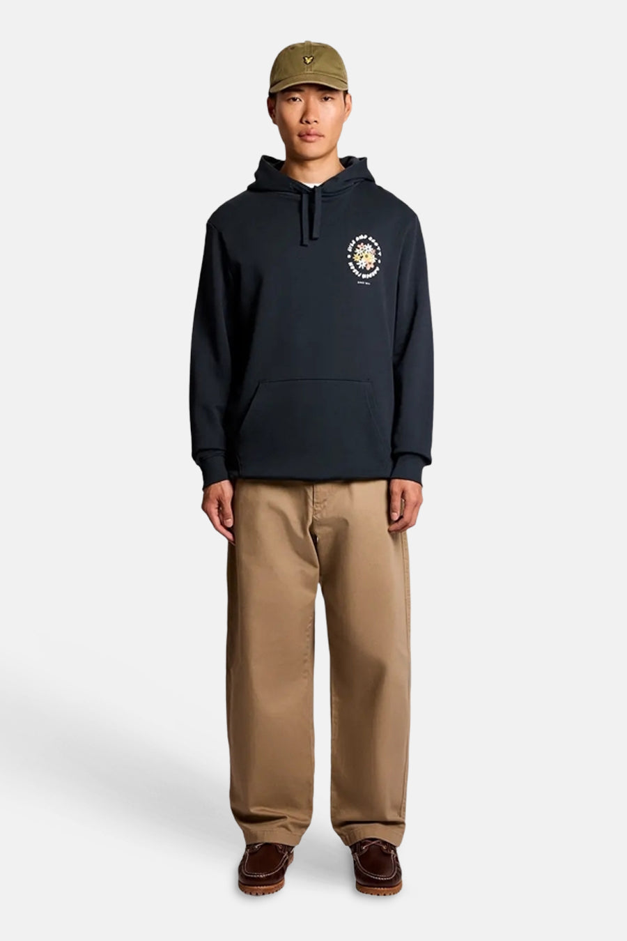 Sweat à capuche bleu de LYLE & SCOTT avec motif floral et texte sur la poitrine, porté avec un pantalon beige et une casquette kaki.