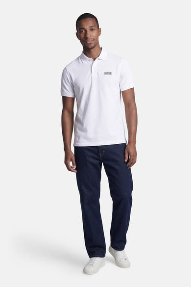 Wit BARBOUR poloshirt met korte mouwen met zwart rechthoekig logo op de borst, gedragen door een man in een spijkerbroek en witte sneakers.