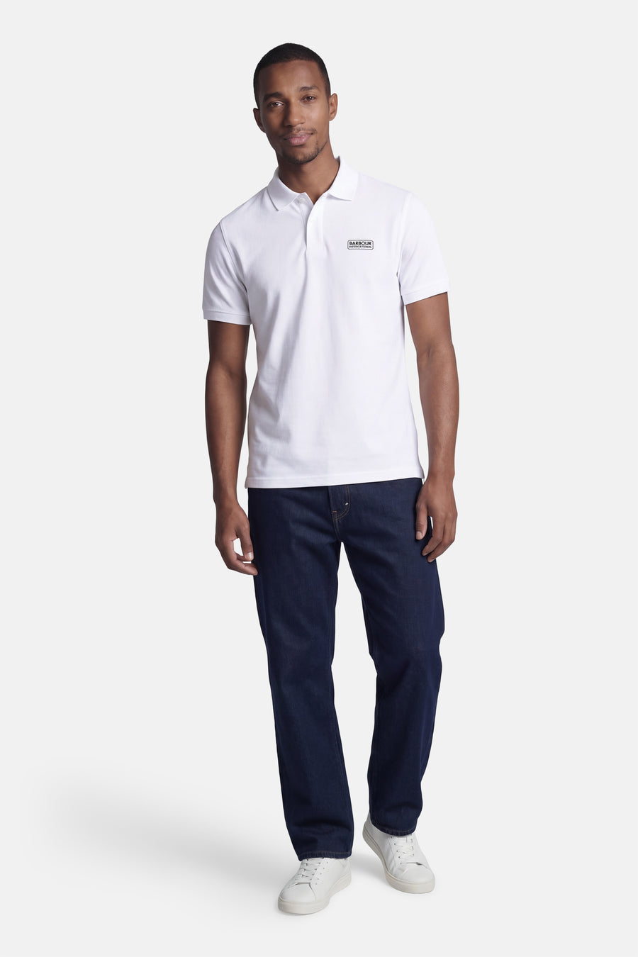 Wit BARBOUR poloshirt met korte mouwen met zwart rechthoekig logo op de borst, gedragen door een man in een spijkerbroek en witte sneakers.