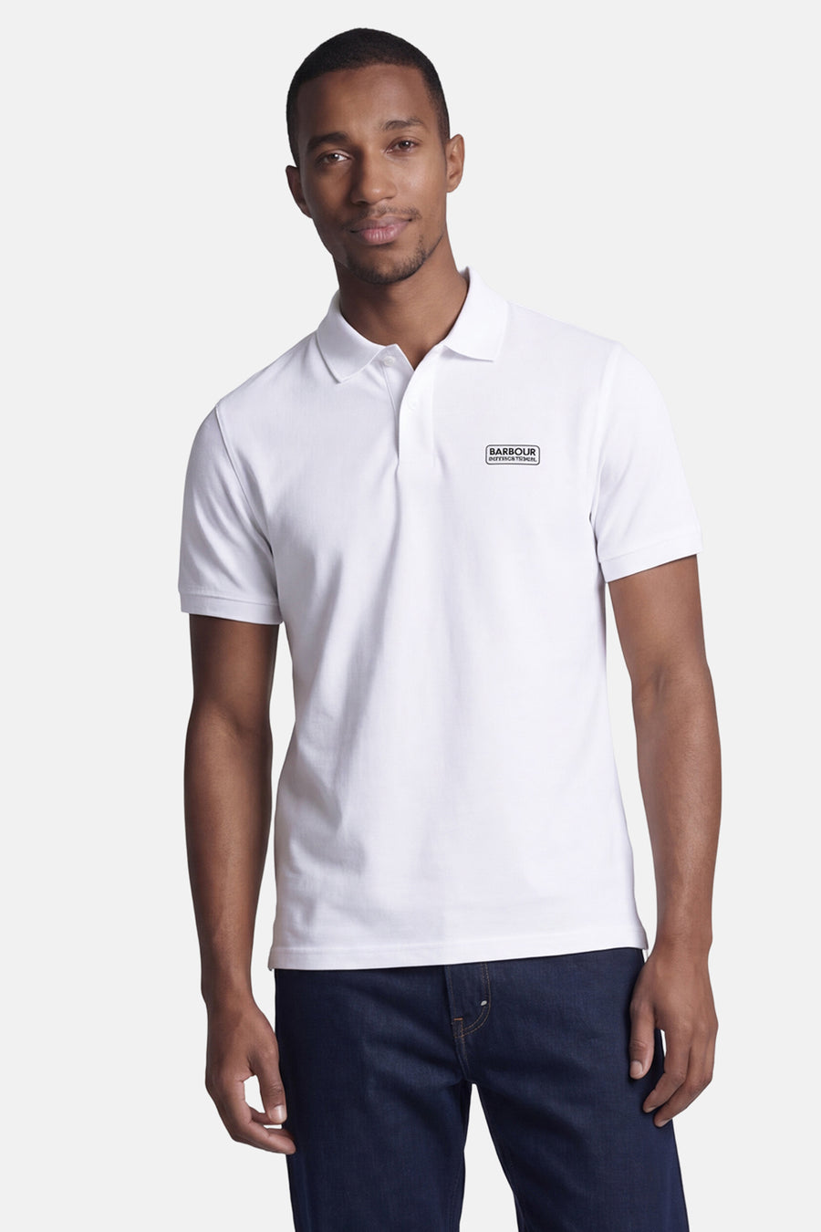Wit BARBOUR poloshirt met korte mouwen, met 'BARBOUR INTERNATIONAL' logo op de borst, gedragen met een donkerblauwe jeans.