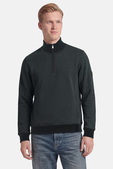 Sweat col montant vert de BARBOUR avec fermeture éclair courte, col côtelé et patch sur la manche, porté avec un jean.