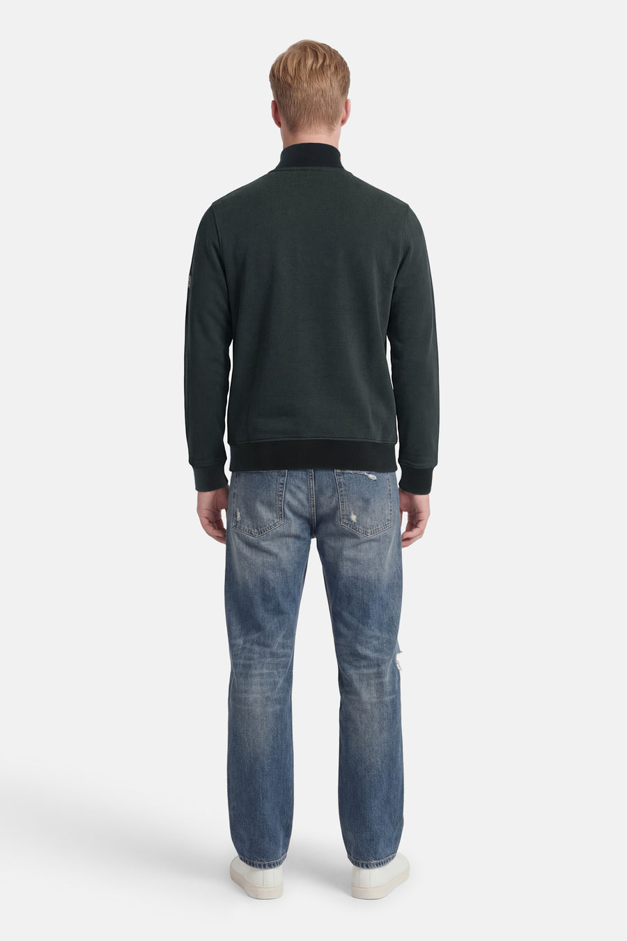 Sweat col montant vert foncé de BARBOUR avec poignets et ourlet côtelés, porté de dos avec un jean déchiré et des baskets blanches.