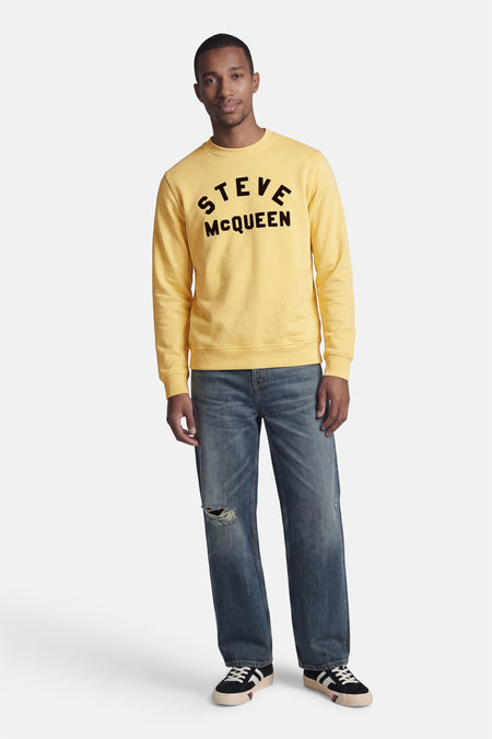 Sweatshirt jaune de BARBOUR avec inscription noire "STEVE McQUEEN", porté par un homme.