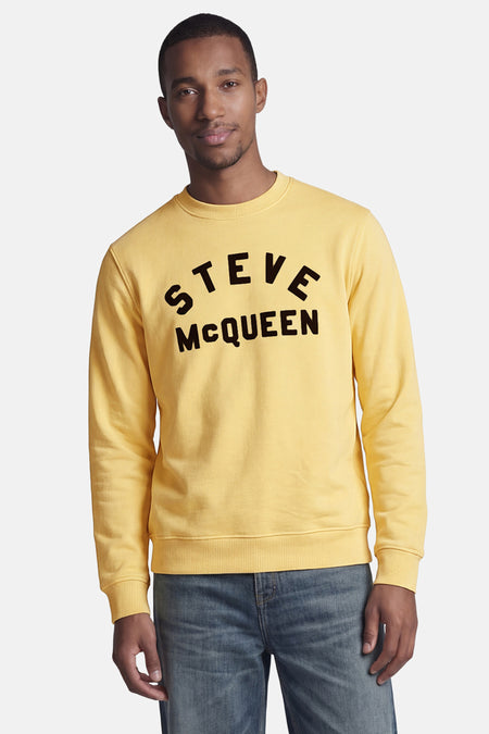 Sweat col rond jaune de BARBOUR avec inscription noire "STEVE McQUEEN" sur un homme en jeans bleus.