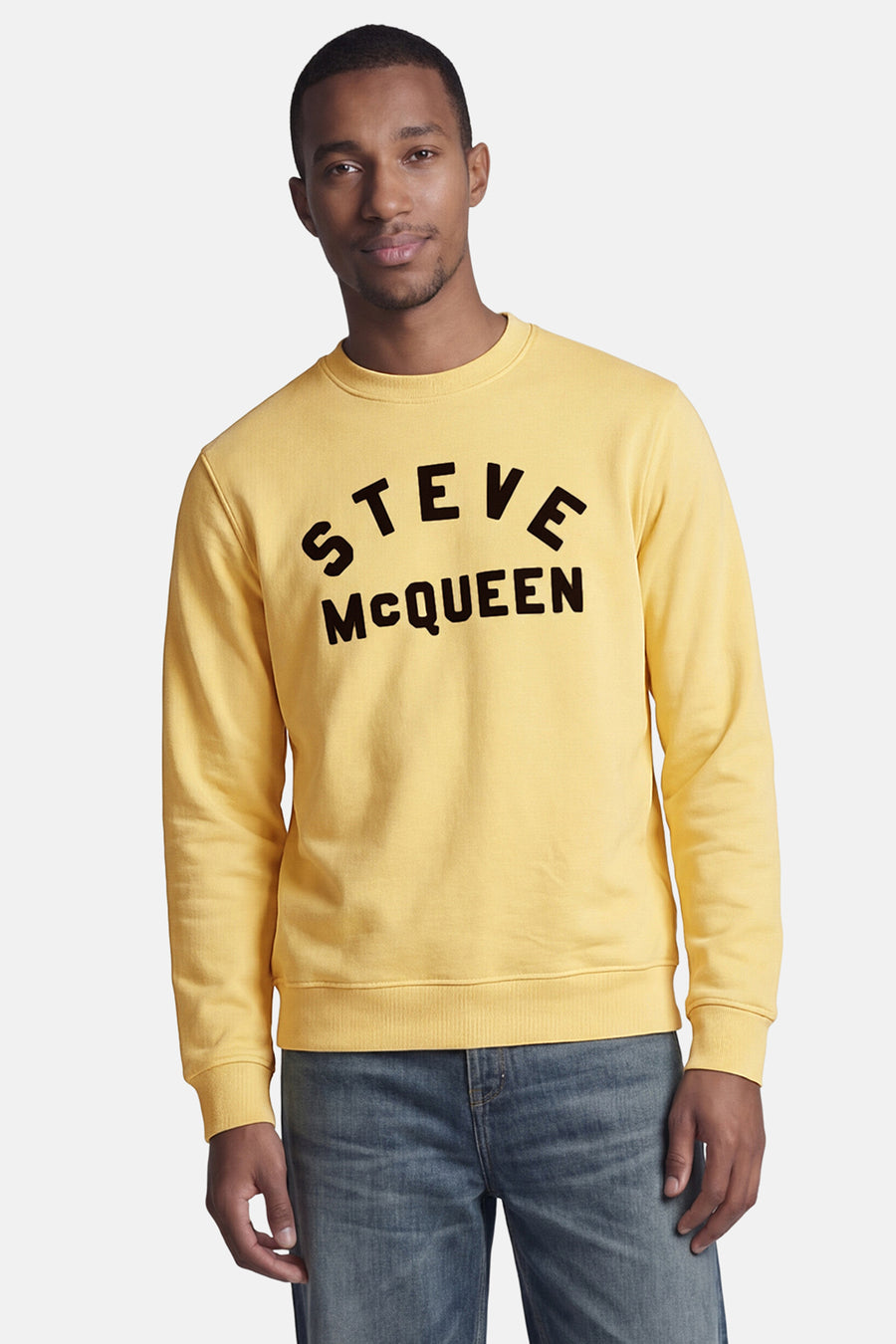 Gele rondehals sweatshirt van BARBOUR met zwarte "STEVE McQUEEN" opdruk op een man in een blauwe jeans.