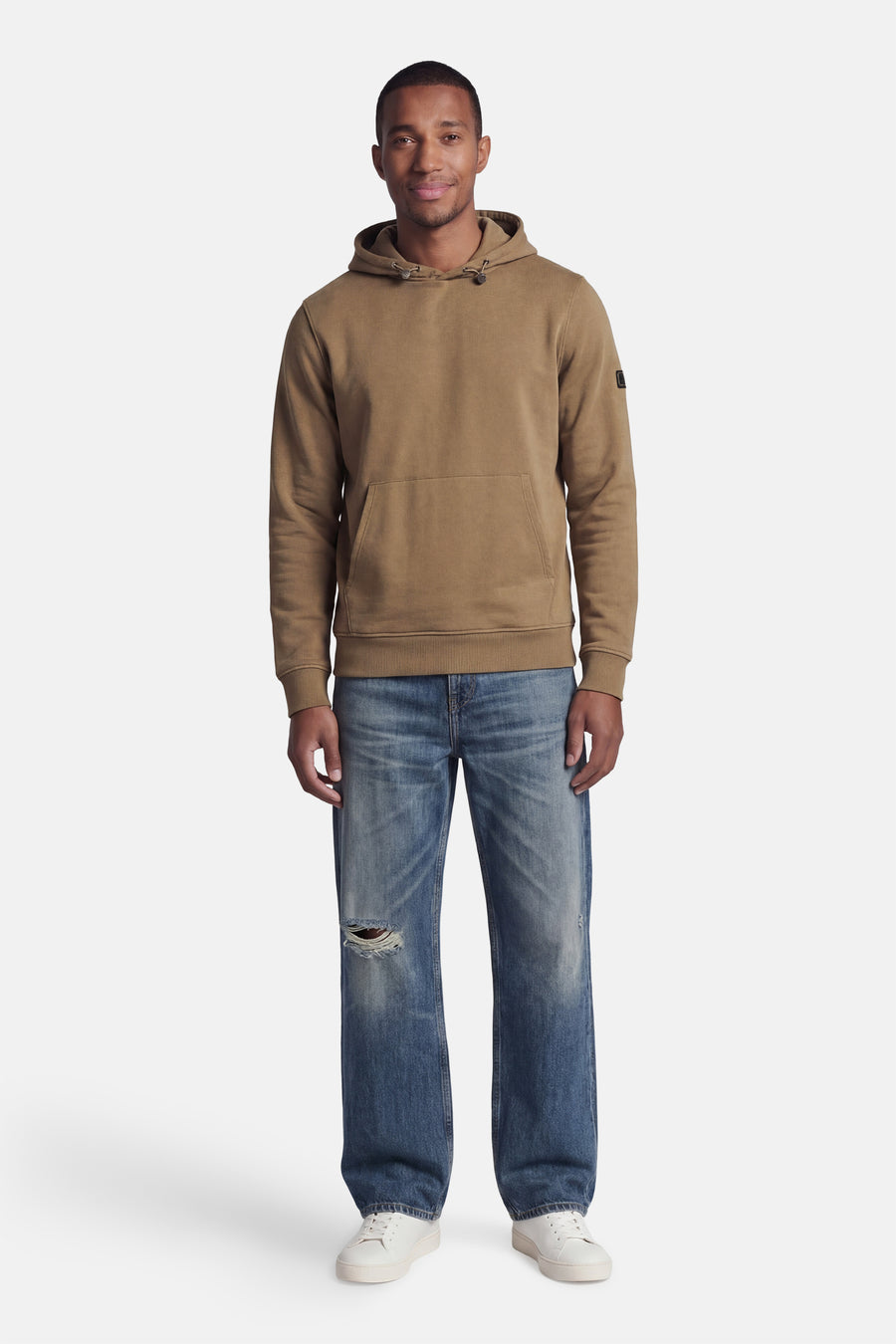 Beige hoodie van BARBOUR met kangoeroezak en trekkoord, gedragen door een man in gescheurde jeans en witte sneakers.