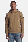 Beige hoodie van BARBOUR met kangoeroezak en zwart embleem op de mouw, gedragen door een man in een blauwe jeans.