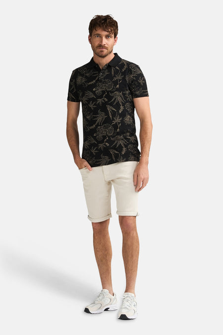 Ecru korte broek van Petrol Industries, gezoomd, gedragen door een man met een zwart poloshirt met bloemenprint en lichte sneakers.
