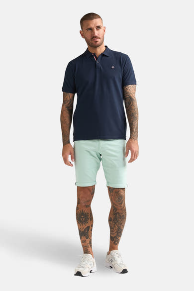 Korte groene short van Petrol Industries, met opgerolde zomen en gedragen met een marineblauw poloshirt en witte sneakers.