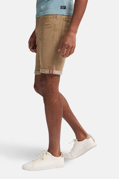 Korte beige short van Petrol Industries, opgerolde zoom met rode bies, gedragen met een blauw T-shirt en witte sneakers.