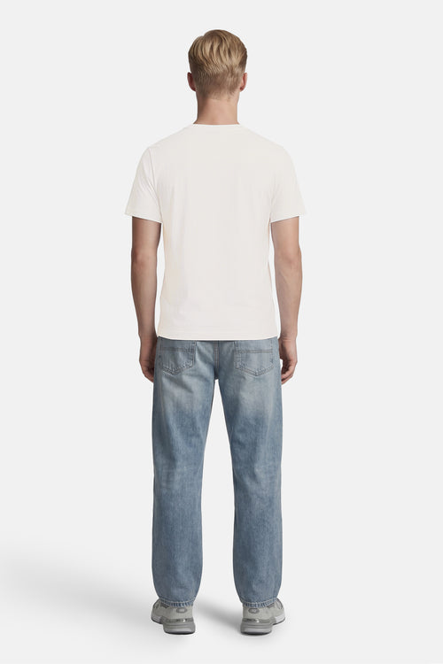 T-shirt blanc à manches courtes de BARBOUR, porté de dos par un homme avec un jean bleu clair et des baskets grises.
