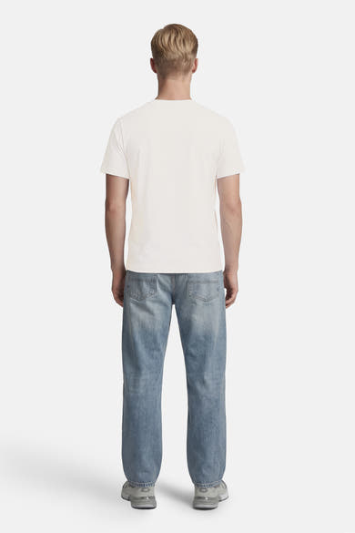 T-shirt blanc à manches courtes de BARBOUR, porté de dos par un homme avec un jean bleu clair et des baskets grises.