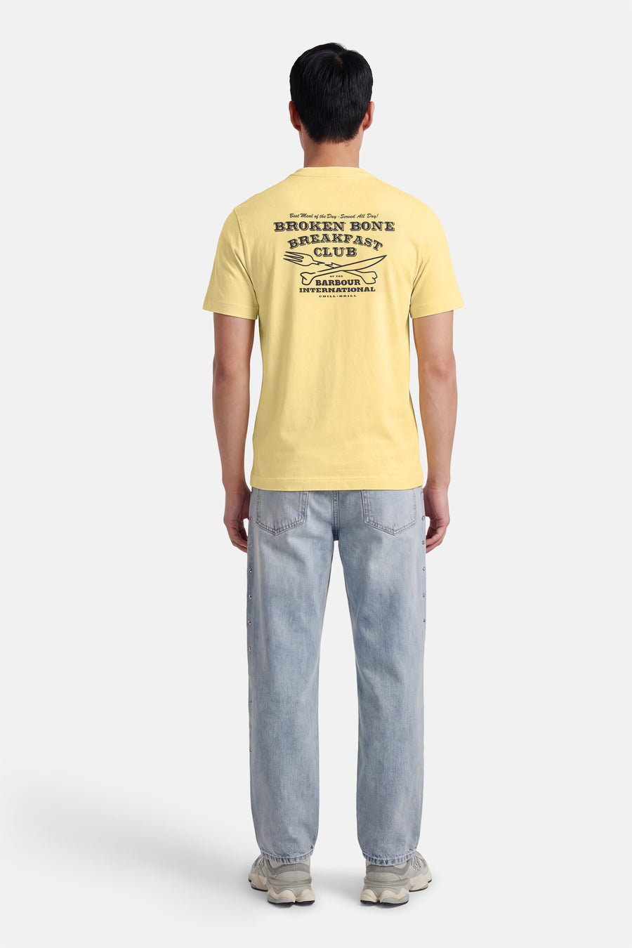 T-shirt à manches courtes jaune de BARBOUR, avec un imprimé graphique au dos, porté avec un jean bleu clair.
