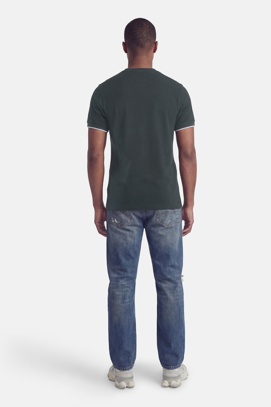 T-shirt à manches courtes vert de BARBOUR, manches soulignées de blanc, porté avec un jean bleu et des chaussures de sport blanches.