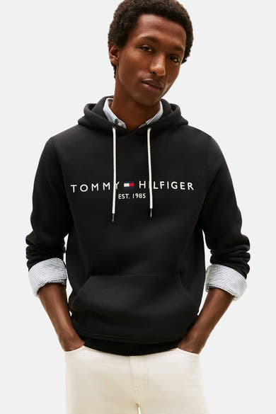 Sweat noir à capuche de TOMMY JEANS, avec cordon de serrage blanc et logo brodé, porté sur une chemise rayée.
