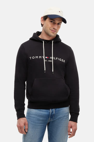 Sweat noir de TOMMY JEANS avec logo Tommy Hilfiger blanc sur la poitrine, capuche à cordons et poche kangourou, porté par un homme en jean et casquette.