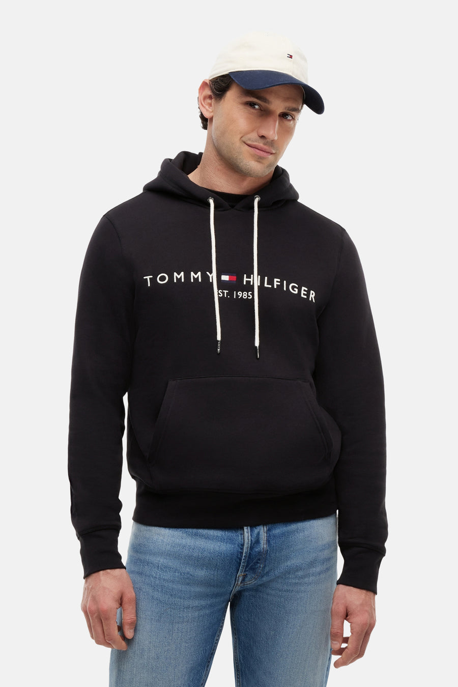 Sweat noir de TOMMY JEANS avec logo Tommy Hilfiger blanc sur la poitrine, capuche à cordons et poche kangourou, porté par un homme en jean et casquette.