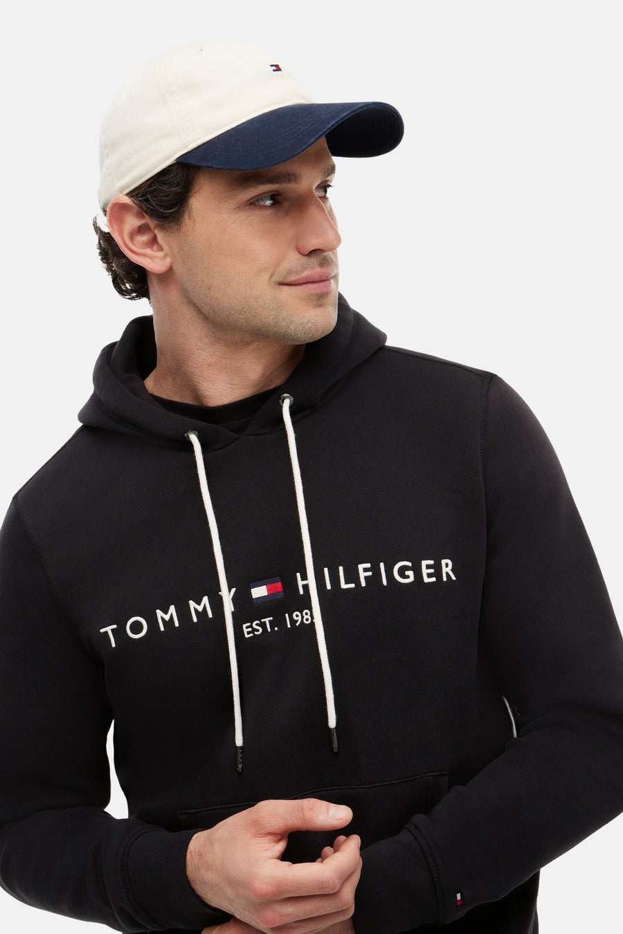 Sweat-shirt noir à capuche de TOMMY JEANS avec cordons blancs, logo Tommy Hilfiger sur la poitrine et un homme en casquette bicolore.