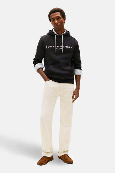 Sweat noir de TOMMY JEANS, porté sur une chemise à rayures et un pantalon couleur crème, avec des chaussures marron.