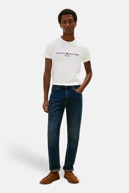 White short-sleeve T-shirt by TOMMY JEANS with chest logo, worn with dark jeans and brown suede shoes, full-body front view.