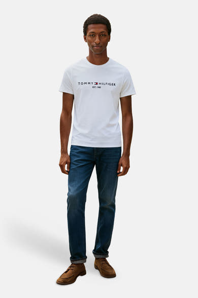 White short-sleeve T-shirt by Tommy Jeans with black logo print, worn with dark jeans and brown loafers, full-body front view