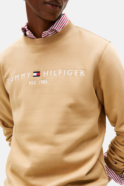 Beige sweater van Tommy Hilfiger met geborduurd logo en ronde hals, gedragen over een gestreept overhemd.