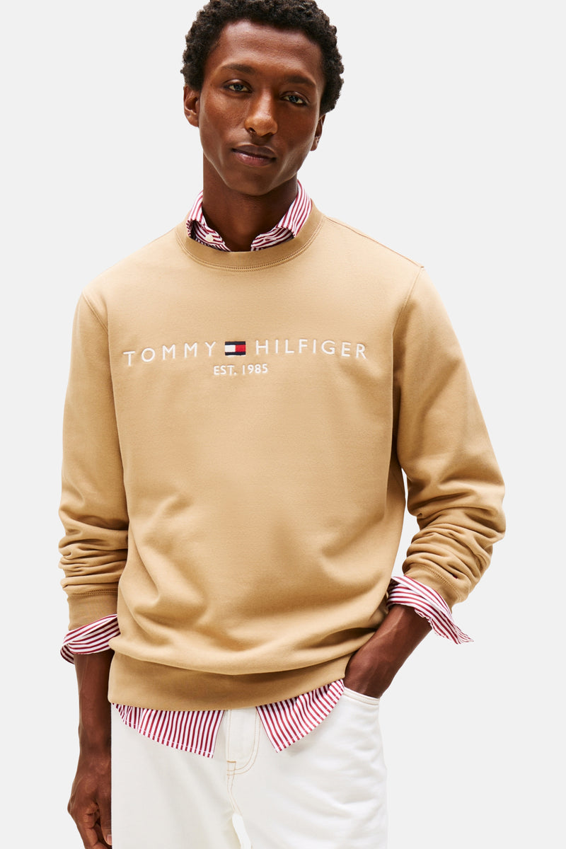 Beige sweater van TOMMY HILFIGER gedragen over een rood-wit gestreept overhemd en een witte broek.